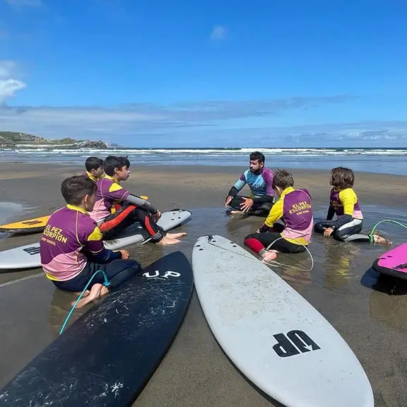 Kinder am Strand mit einem Coach, der vor einer Surfstunde im internationalen Sommerlager in Spanien erklärt