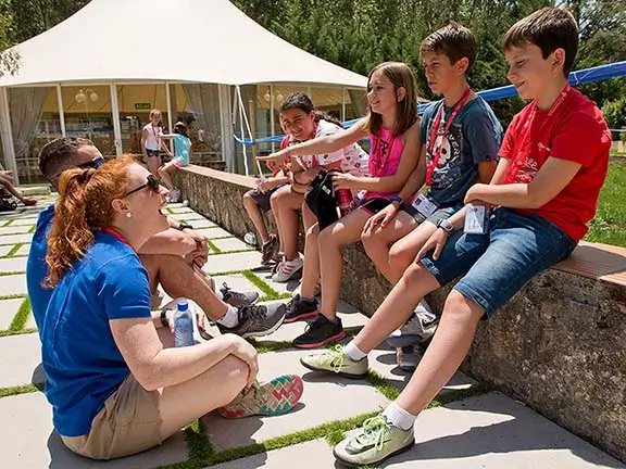 Fünf Kinder unterhalten sich mit zwei internationalen Coaches in einem Gartenbereich während eines internationalen Sommerlagers in Spanien.