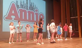 Children in a summer camp rehearsing a musical with professional English-speaking actors, singers, and dancers.