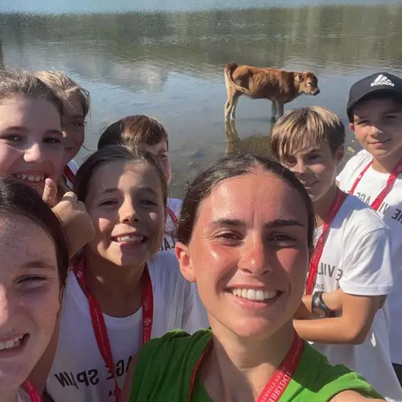 A group of kids with an international coach by a lake with a cow during an excursion