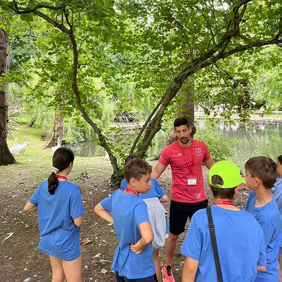 Un coach internacional habla con un grupo de chicos frente a un lago en una excursión del campamento de inglés